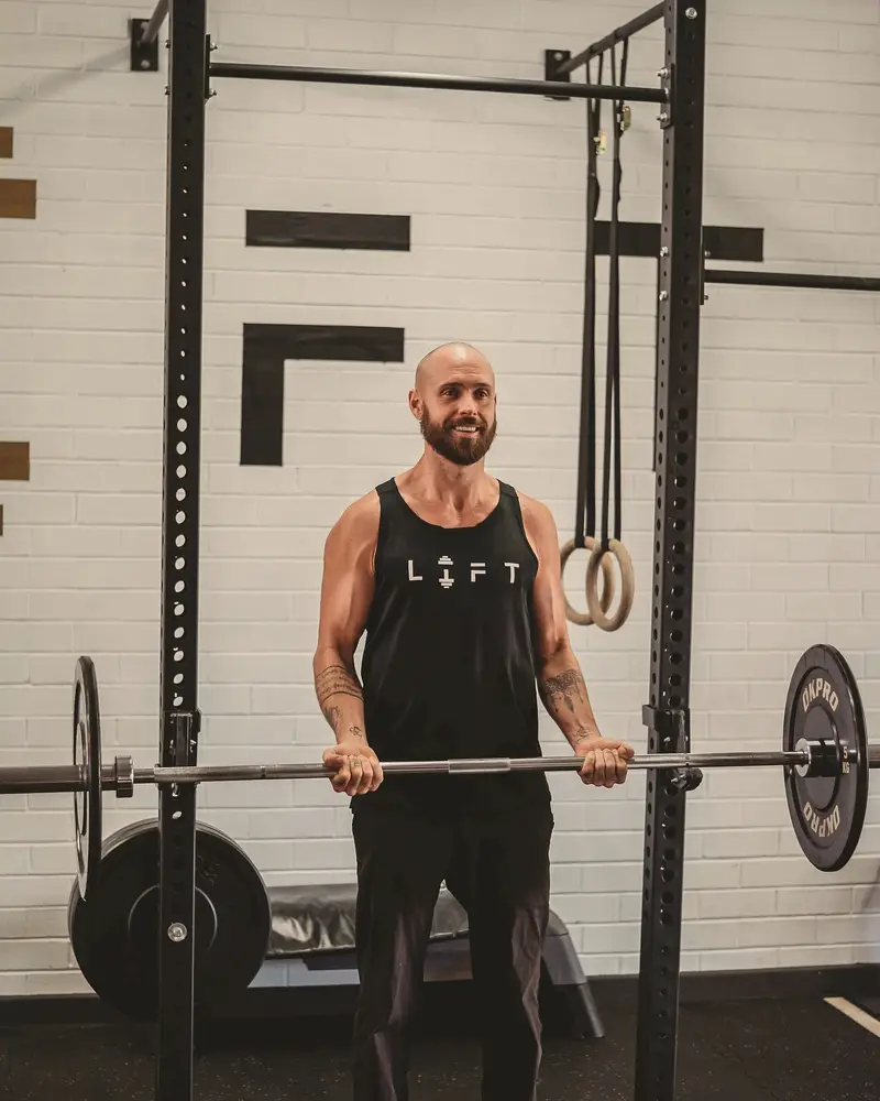 Coach Chris smiles while holding a barbell at hip height inside a squat rack.