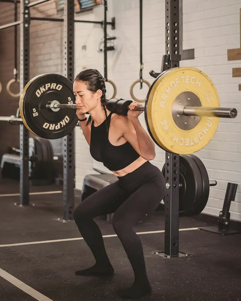 Coach Lisa sits into a back squat with a loaded barbell in a squat rack.