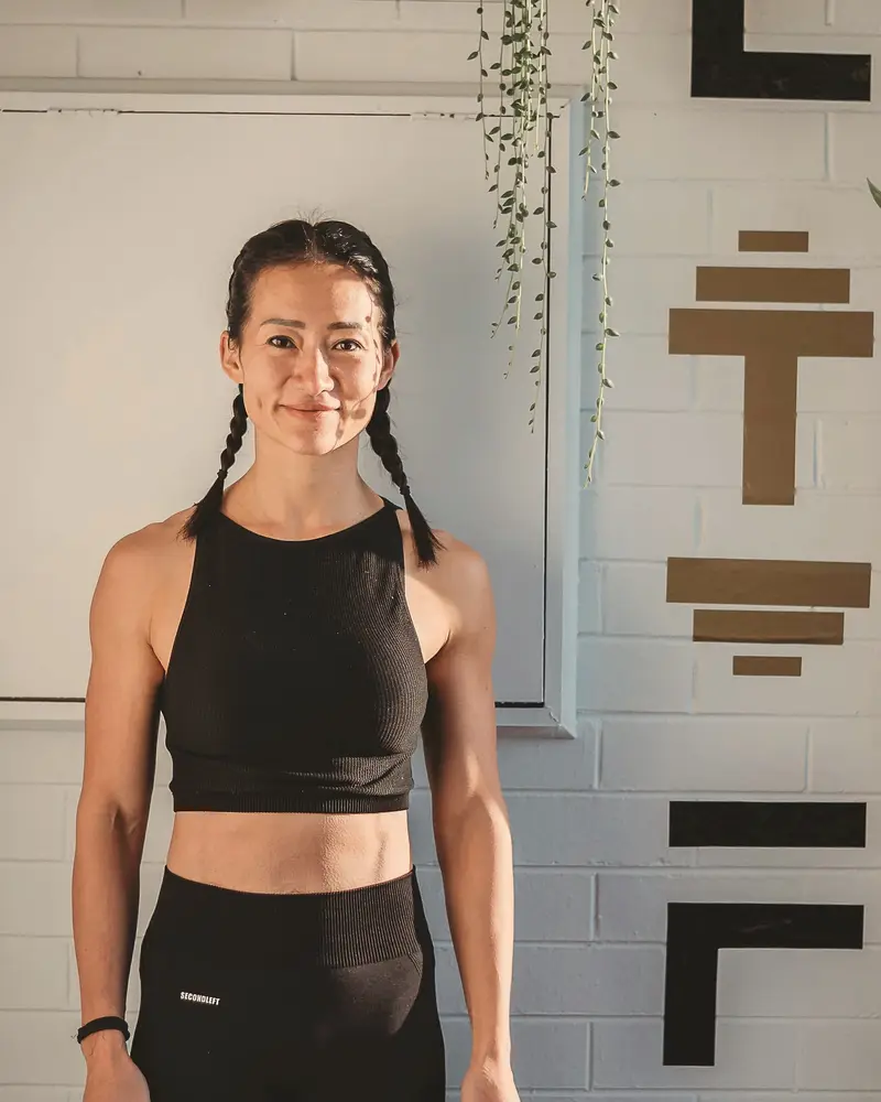 Coach Lisa, in black training clothes with her hair in two plaits, stands beside the LIFT lettering on a white brick wall.