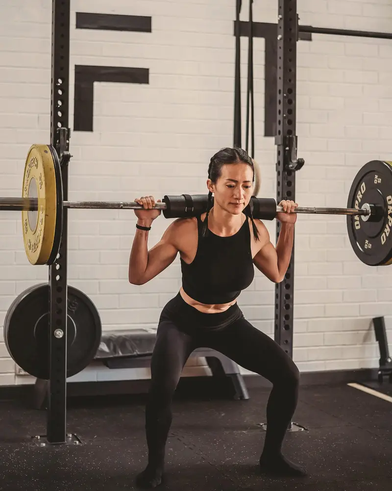 Coach Lisa squats with a padded barbell across her shoulders inside a squat rack.
