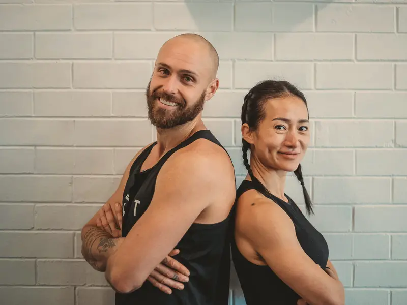 Coaches Chris and Lisa stand back to back with their arms crossed, smiling, against a white brick wall.