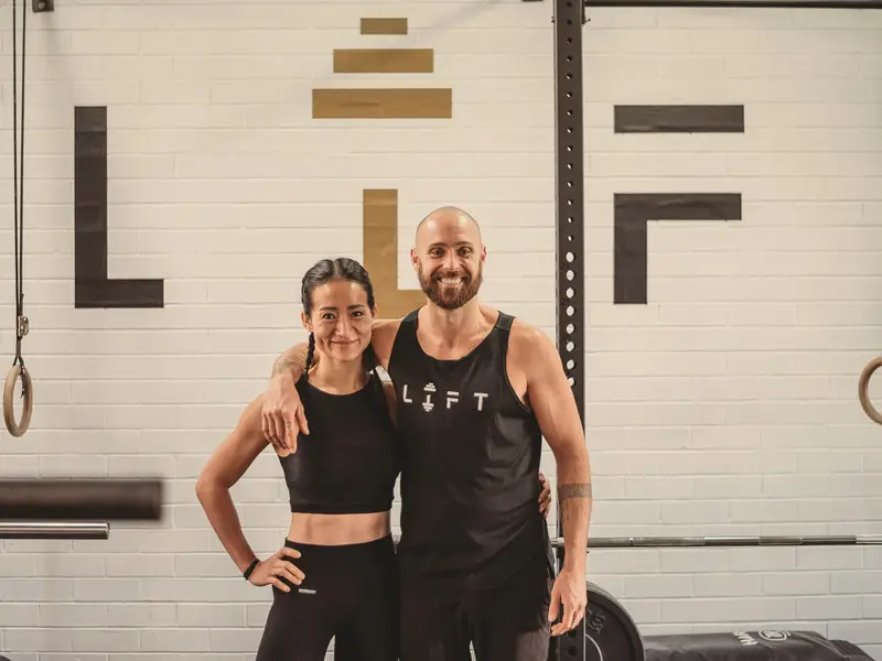 Coaches Chris and Lisa smile with their arms around each other in front of the black and gold LIFT logo, with gymnastic rings on either side.