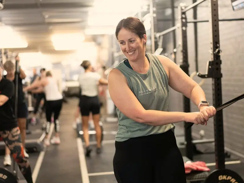 A smiling woman pulls a resistance band across her body in a class, with members training along the racks behind her.
