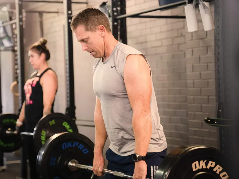 A man in a grey singlet focuses as he lifts a barbell in a class, with another member lifting behind him.