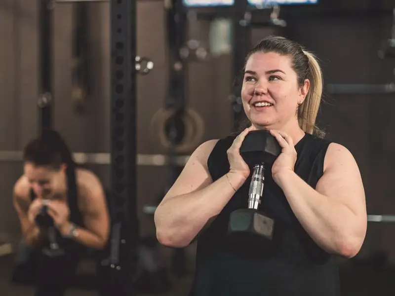 A smiling woman holds a dumbbell at her chest for a goblet squat, with another member training behind her.