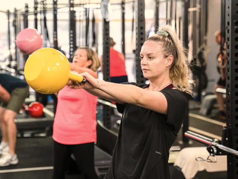 A woman swings a yellow kettlebell in a busy class, with other members working at the racks behind her.