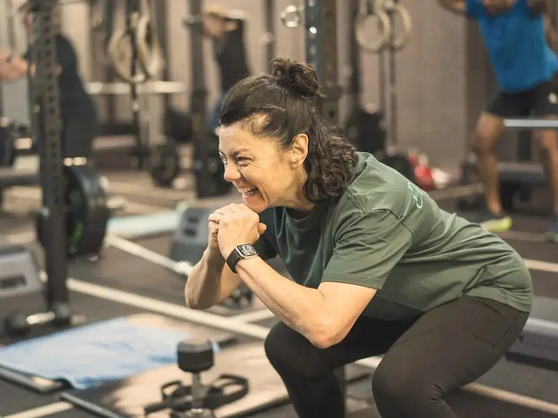 A woman in a green T-shirt laughs while holding a squat during a class, with other members training behind her.