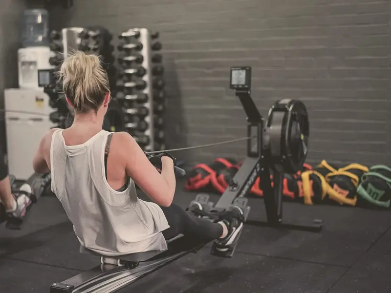 A woman rows on a rowing machine, seen from behind, with dumbbell racks and sandbags in front of her.