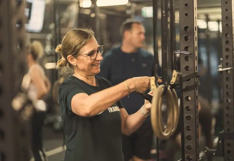 A smiling woman in glasses adjusts the straps of a set of gymnastic rings on a rack during a session.
