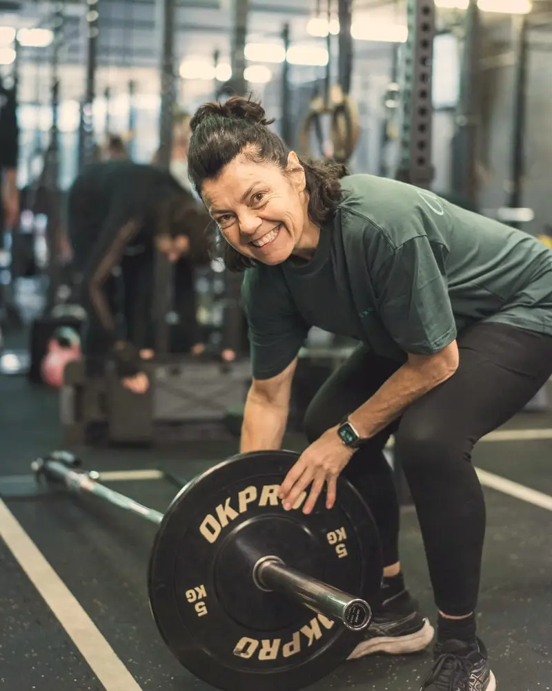 A smiling woman in a green T-shirt bends to set up a barbell loaded with small plates, with a class training behind her.