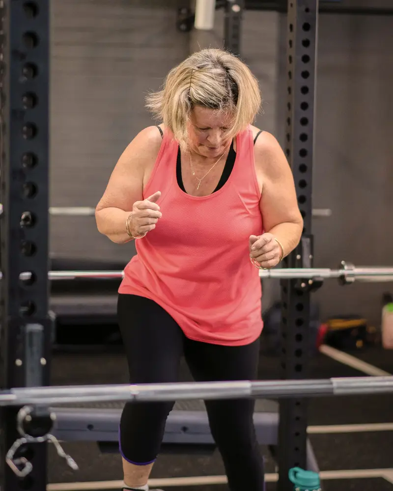 A woman in a coral singlet steps up onto a low step between two squat racks during a class.
