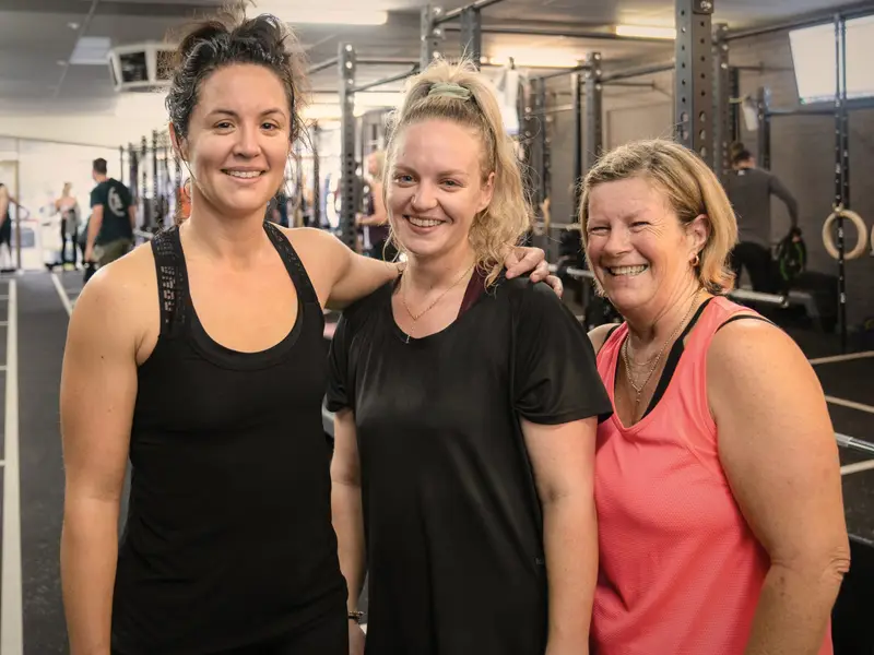Three women of different ages stand together smiling on the gym floor after a class.
