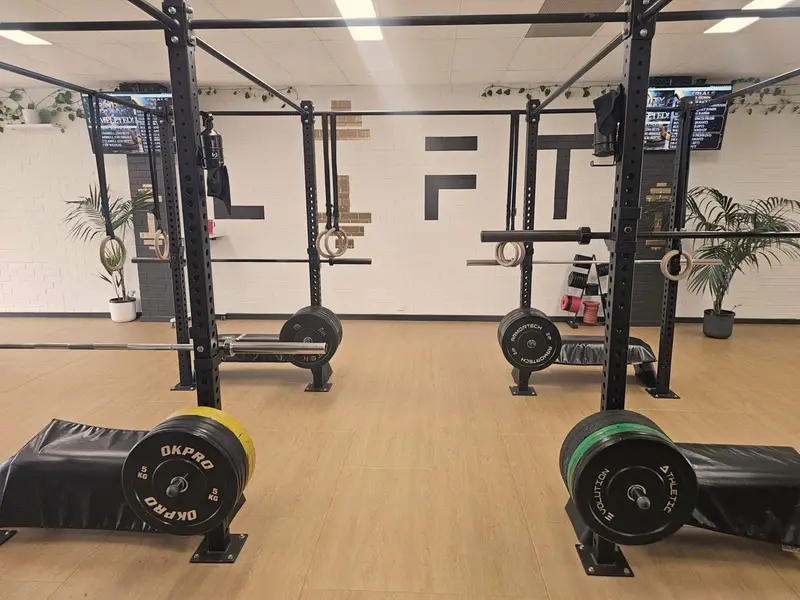 LIFT Gym Wangara training floor: squat racks with gymnastic rings and loaded barbells on a timber floor, in front of the white brick wall with the black LIFT lettering.
