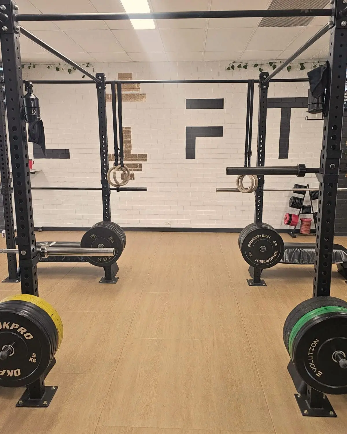 LIFT Gym Wangara training floor: squat racks with gymnastic rings and loaded barbells on a timber floor, in front of the white brick wall with the black LIFT lettering.