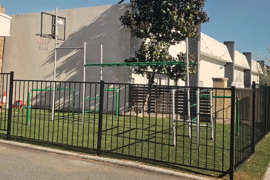 The fenced outdoor playground beside the gym: artificial grass, a basketball hoop and climbing bars behind a black metal fence.