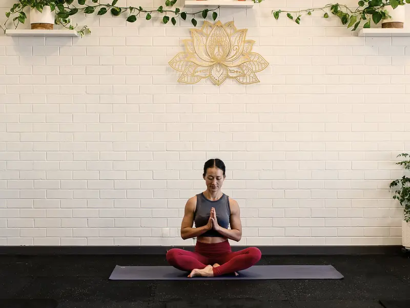 Coach Lisa sits cross-legged on a mat with her hands together, under a gold lotus emblem and trailing plants on a white brick wall.